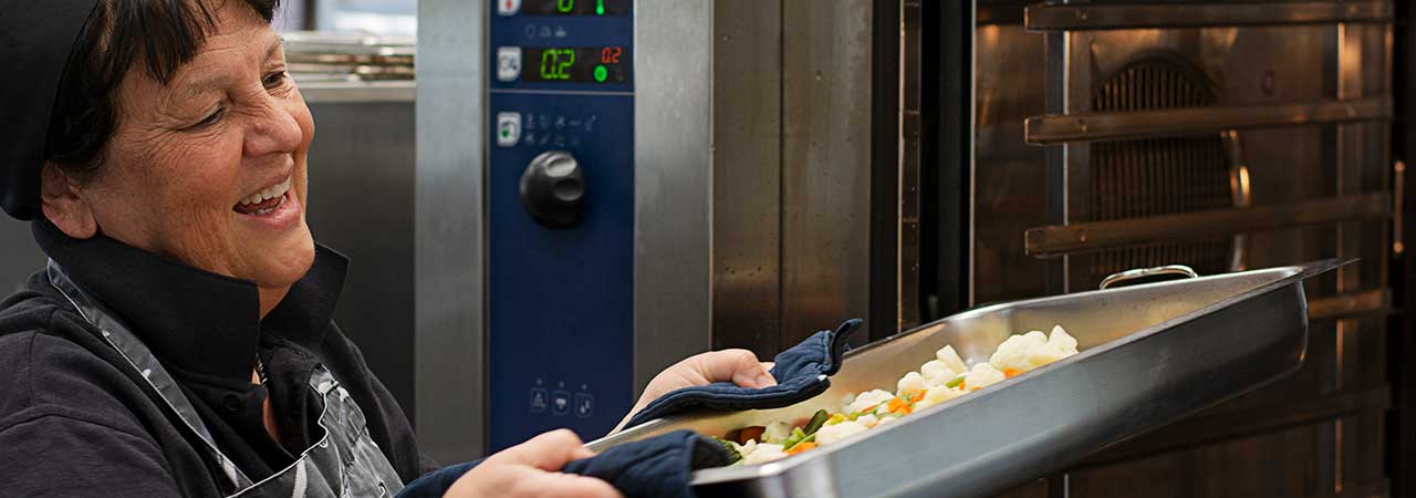 Kvinna i skolkök tar ut mat ur ugn.